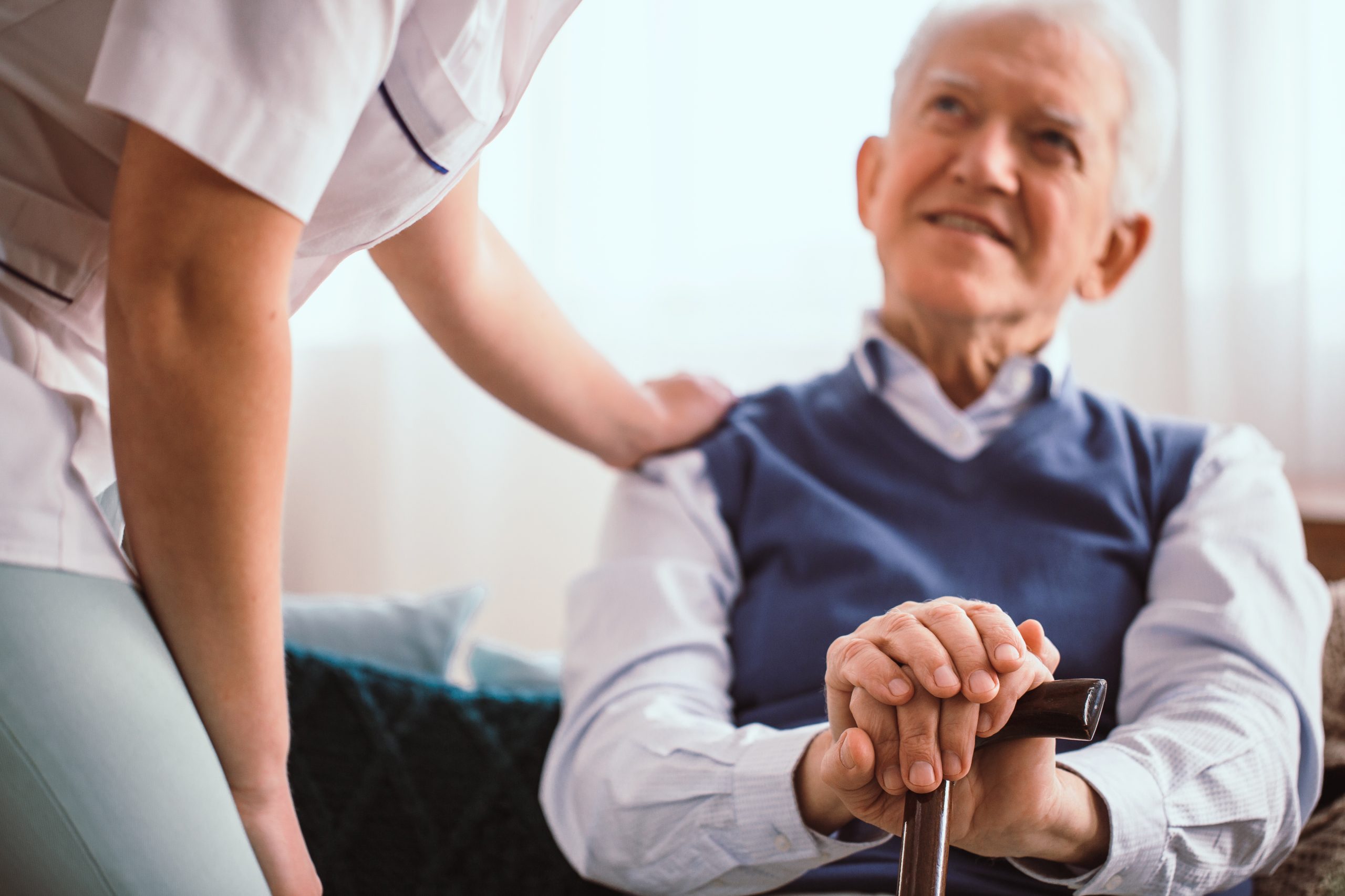 Primer plano de un anciano sonriente que apoya las manos en un bastón, con la mano de un cuidador colocada suavemente sobre su hombro, simbolizando consuelo y apoyo.