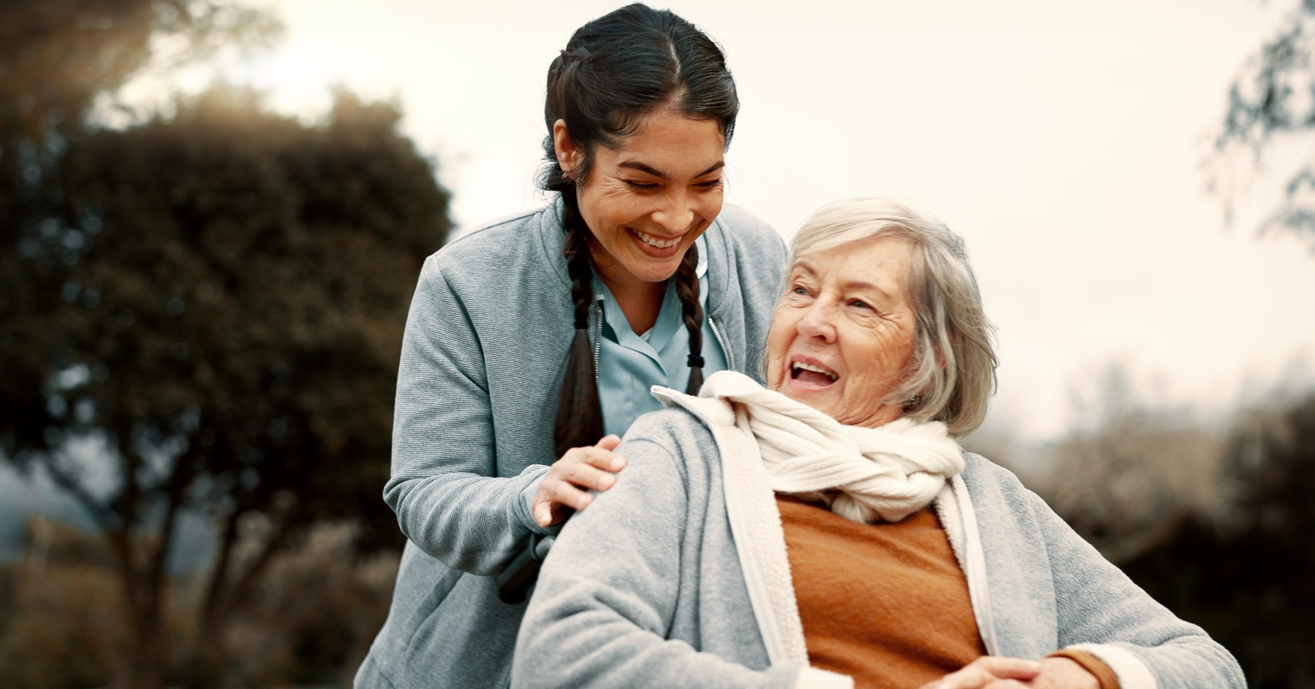 Una joven cuidadora sonriente con uniforme de enfermera empuja la silla de ruedas de una anciana feliz al aire libre.