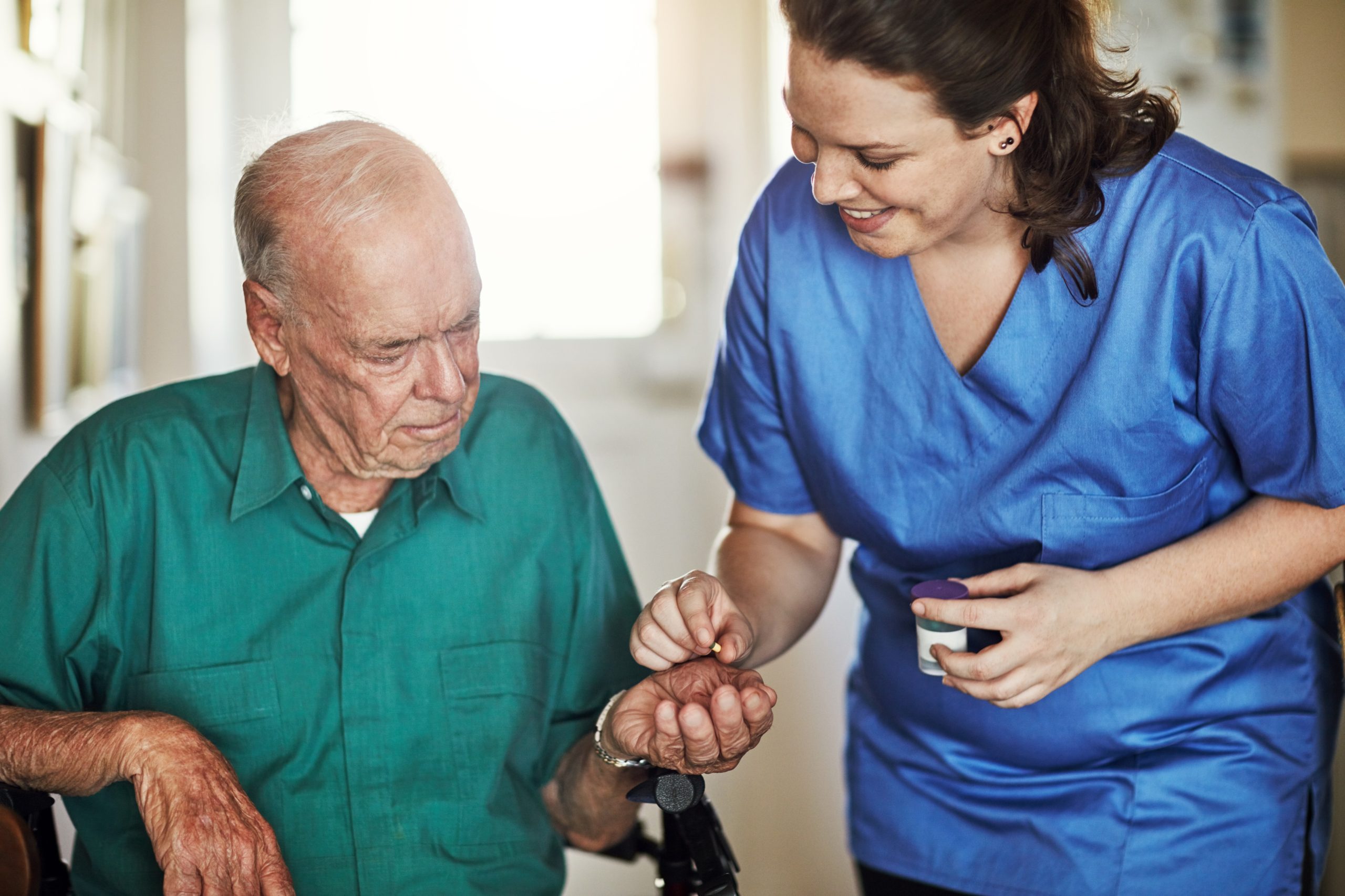 Una enfermera sonriente, vestida con uniforme azul, administra medicación (una pequeña pastilla) a un anciano sentado en una silla, que mira la pastilla en la palma de su mano.