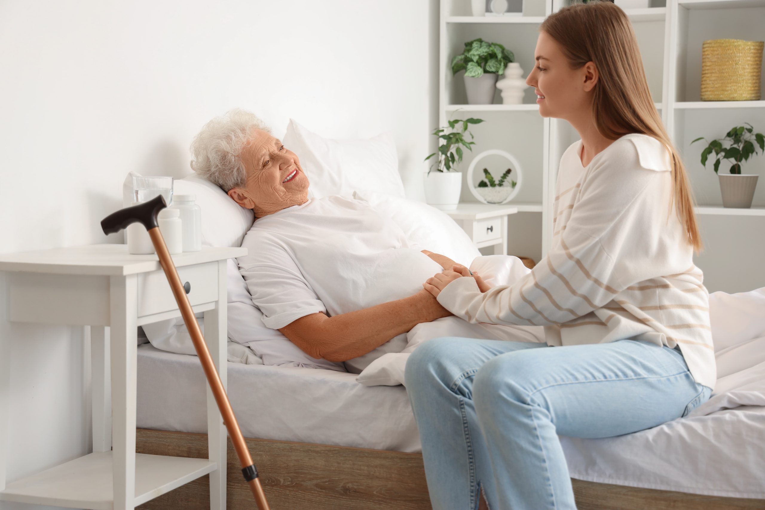 Una joven sentada junto a la cama de una anciana sonriente en una habitación luminosa, le toma la mano para consolarla y hacerle compañía. Un bastón descansa sobre una mesilla cercana.